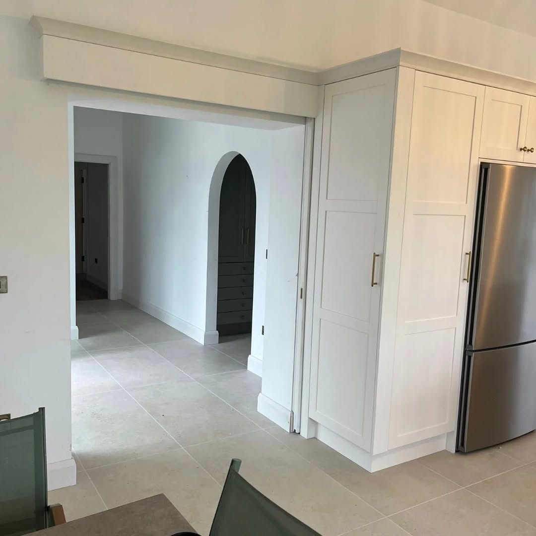 White fitted kitchen with fridge wall viewed through an arched opening.